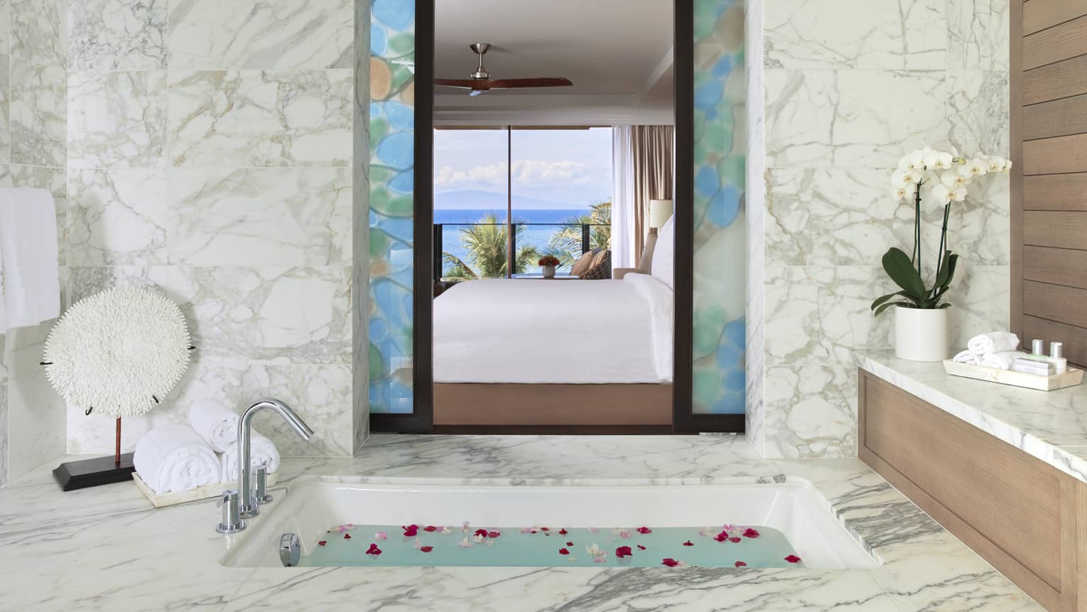Luxurious bathroom with marble accents, a soaking tub with rose petals and a view of the bedroom and ocean through decorative glass panels