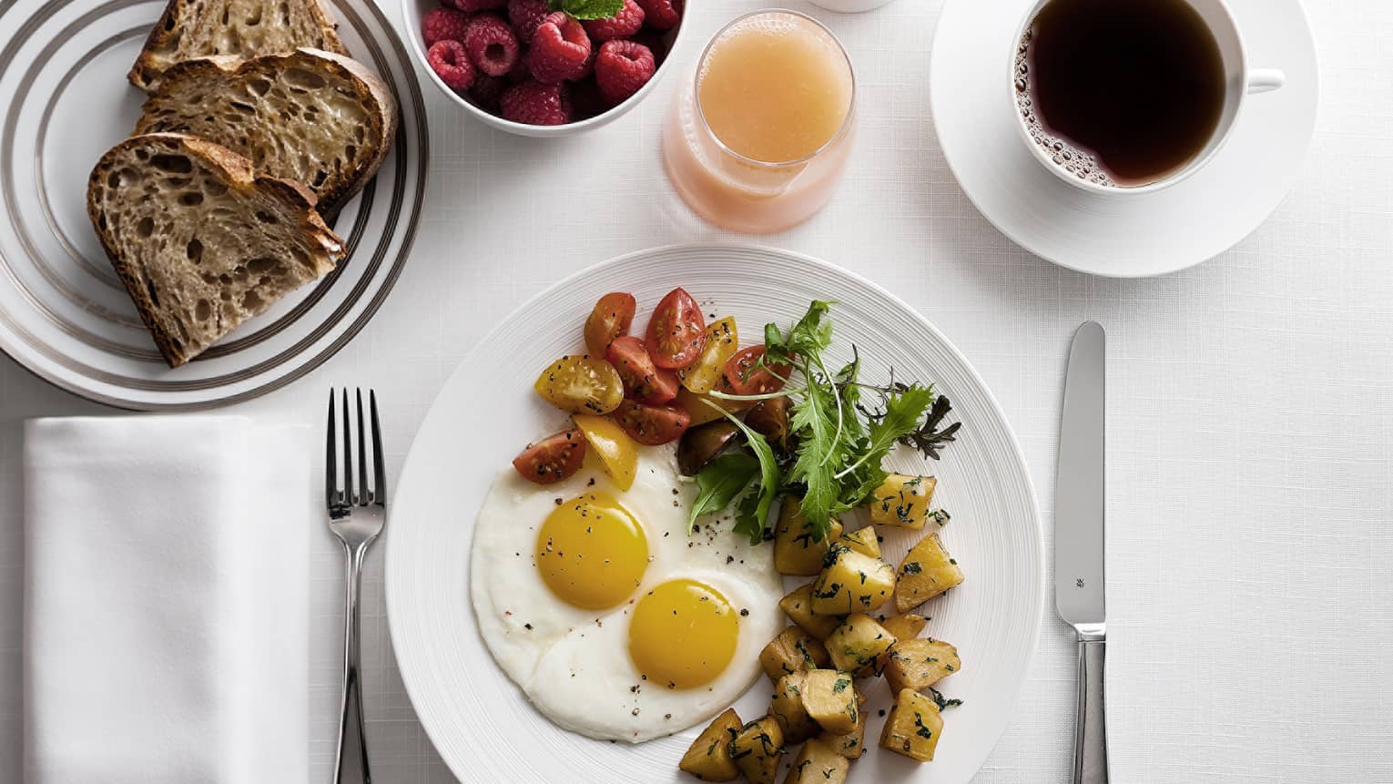 Breakfast plate with sunny-side-up eggs, cherry tomatoes, greens, and roasted potatoes, alongside toast, raspberries, grapefruit juice and coffee.