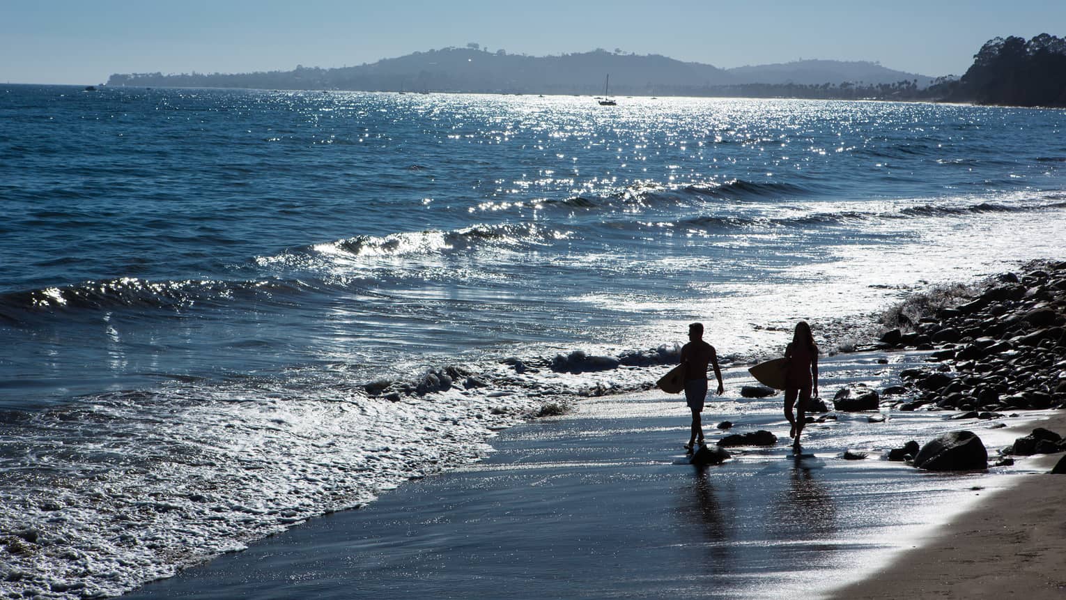 Silhouettes of two people carrying surfboards along beach shoreline