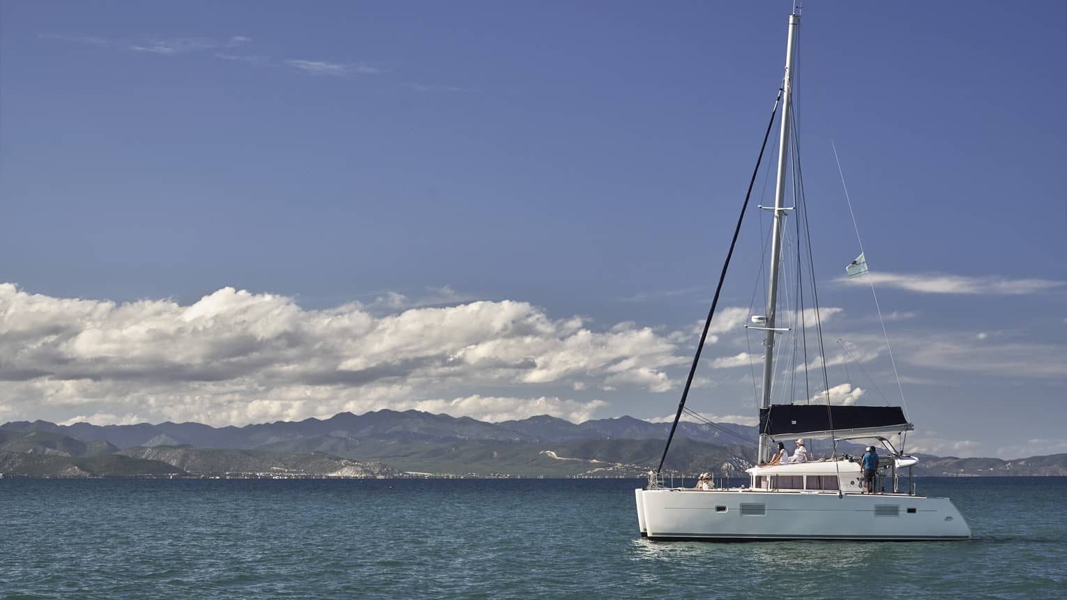 A catamaran on calm, blue water under a partly-cloudy sky with low green hills in the background and mountain ranges beyond.