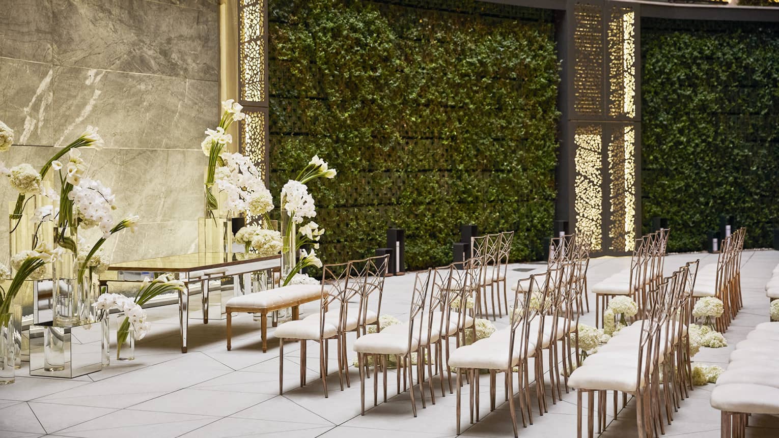 Five rows of white-and-gold chairs set up facing an antique table and bench surrounded by large white floral arrangements, all set in an intertior courtyard featuring curved, vine-filled walls