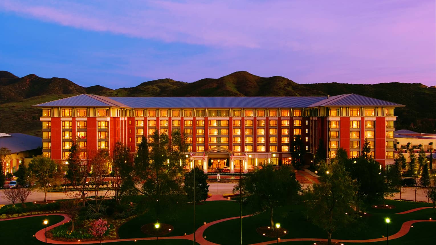 The exterior of a large hotel building surrounded by trees and mountains, the sun is setting in the background and the sky is pink and blue.