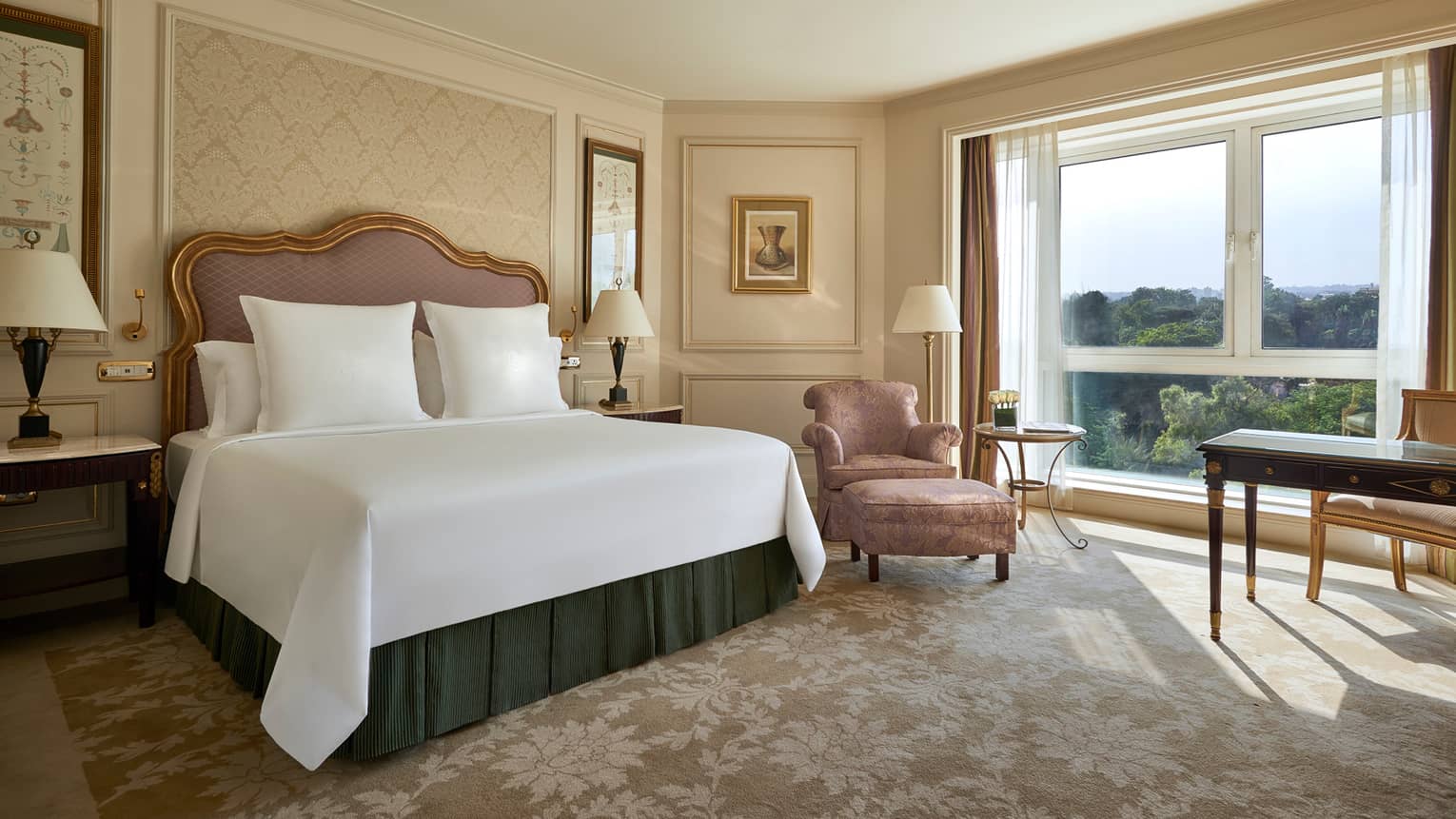 Deluxe Room bed with upholstered pink headboard, gold pattern wallpaper, pink silk armchair by sunny window