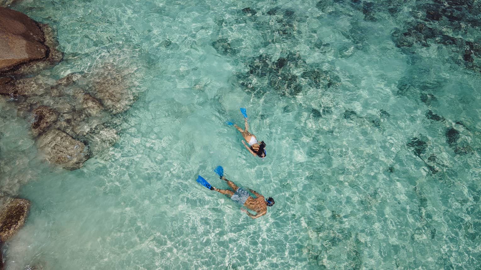 Aerial view of two snorkellers wearing blue flippers in an expanse of shallow, turquoise waters rippling over the reef.