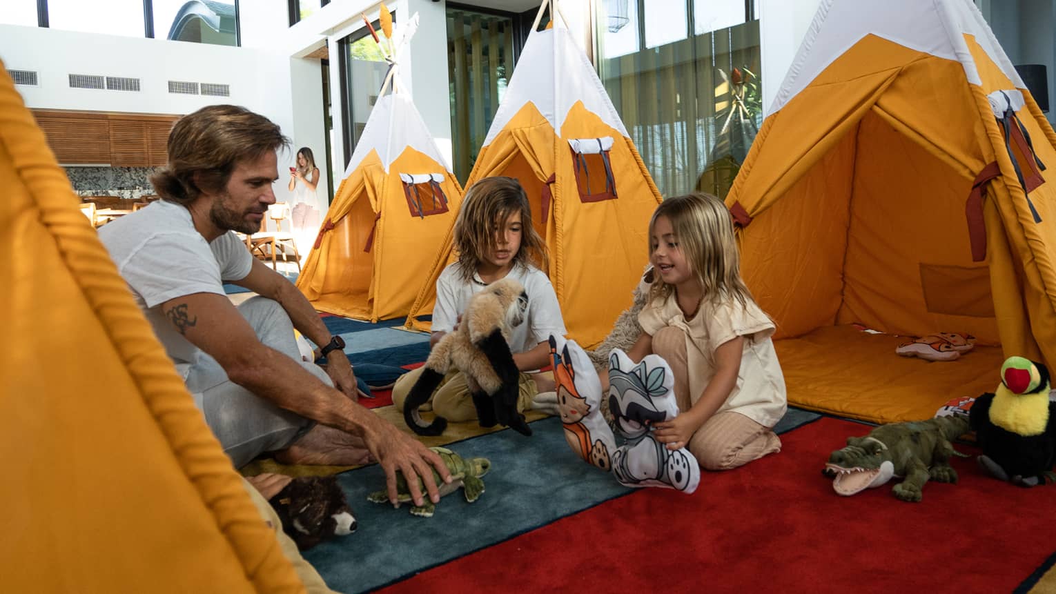 Two young kids and an adult play with stuffed animals near a row of orange indoor play tents