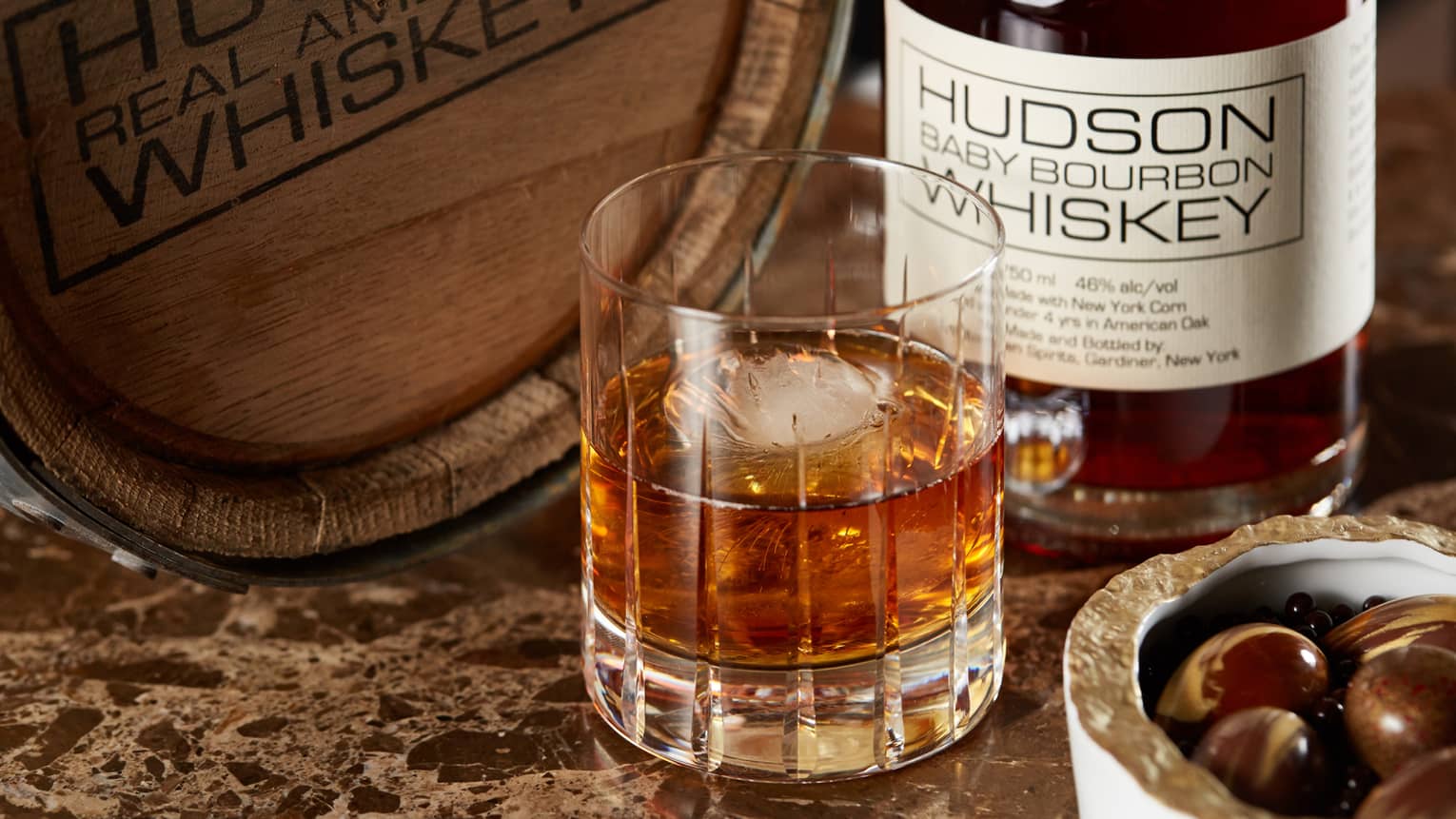 Whiskey with ice in crystal glass on table by Hudson Baby Bourbon Whiskey bottle, barrel