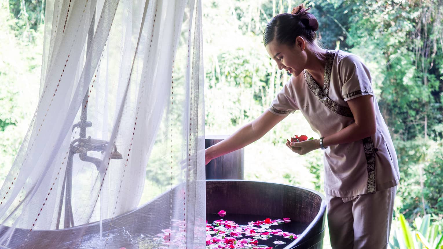 Hotel staff drops colourful flower petals in free-standing copper bathtub draped with sheer white net 