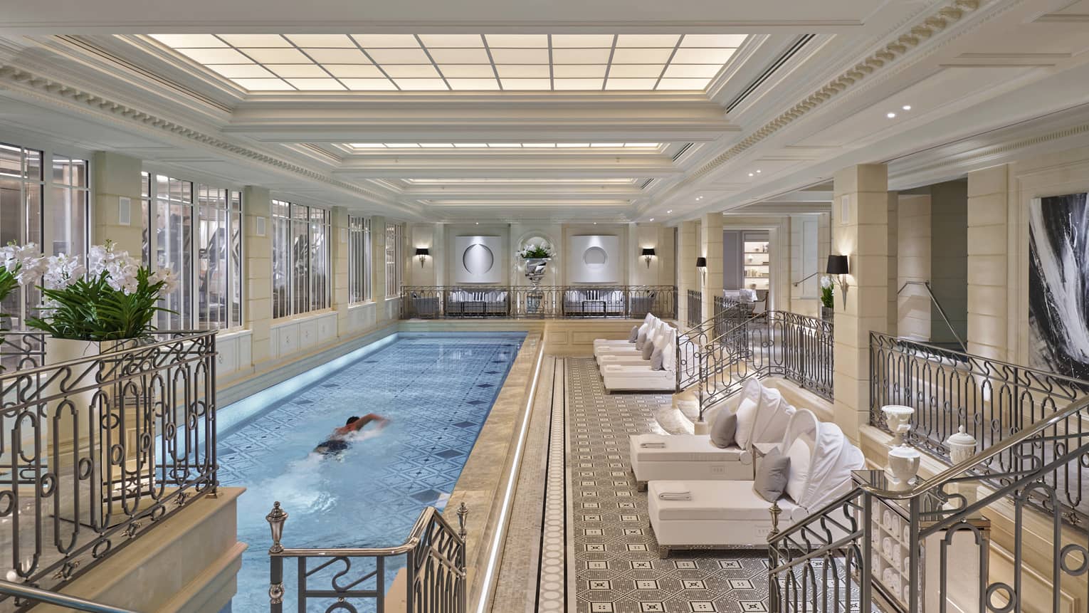 A man swims in an indoor, long, rectangular pool with pool deck featuring white lounges
