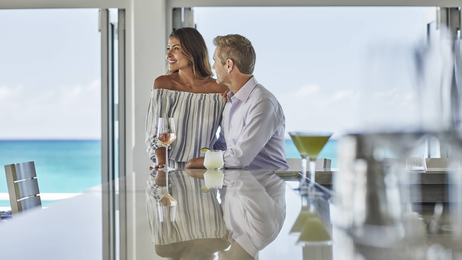 Couple with cocktails at end of shining bar by bright windows overlooking ocean