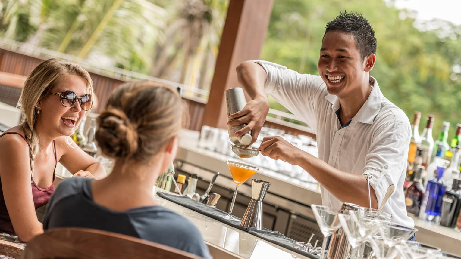 Smiling mixologist pours orange cocktail in martini class by two women at bar