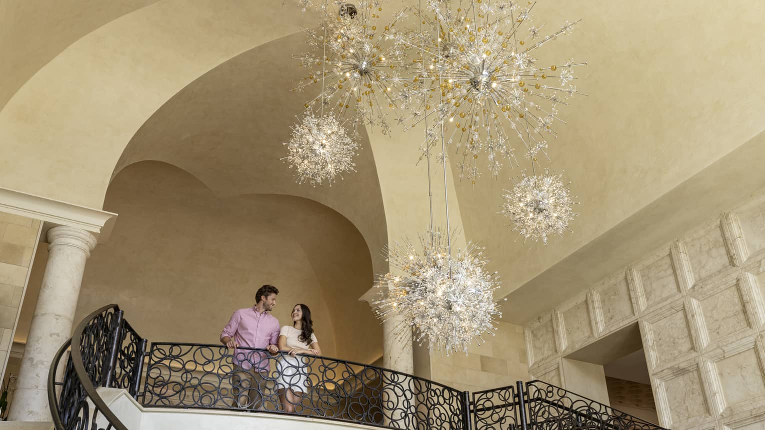 A man and woman standing at the top of a stairway with an open space looking at a large chandelier.