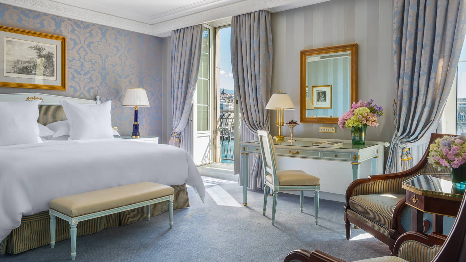 Elegant hotel room with a king bed, desk, seating area and a balcony overlooking water, featuring light blue and gold decor