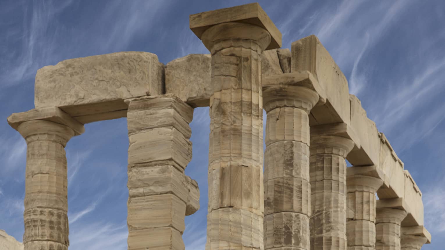 Side view of ancient ruins with formidable Doric columns on a stone foundation, cloud-streaked sky and hilly islands beyond.