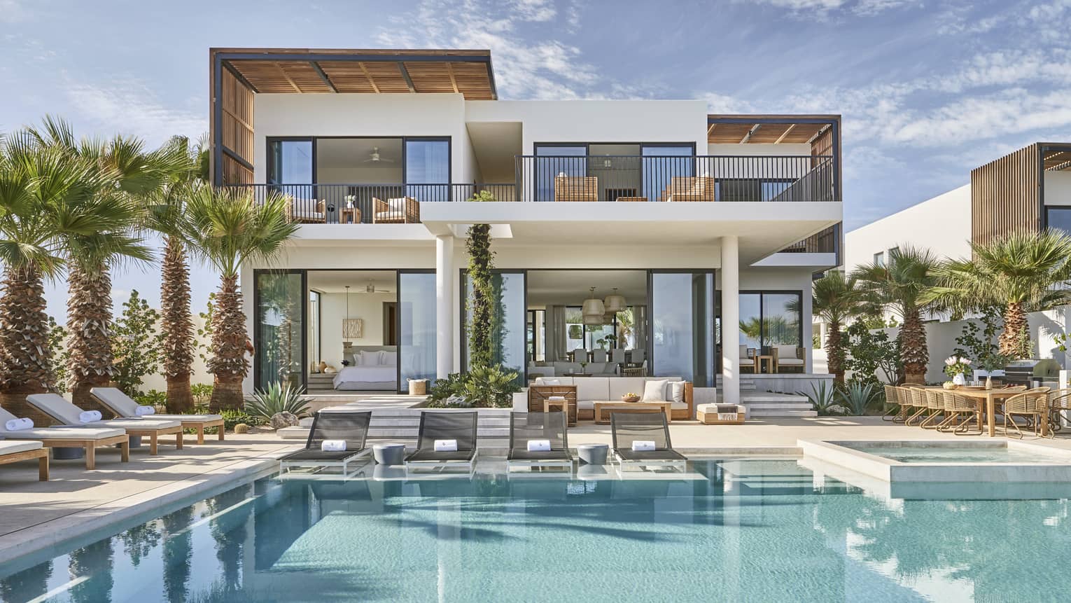 Modern luxury villa with a spacious pool area, surrounded by palm trees, featuring outdoor lounge chairs, a dining area, and floor-to-ceiling windows.