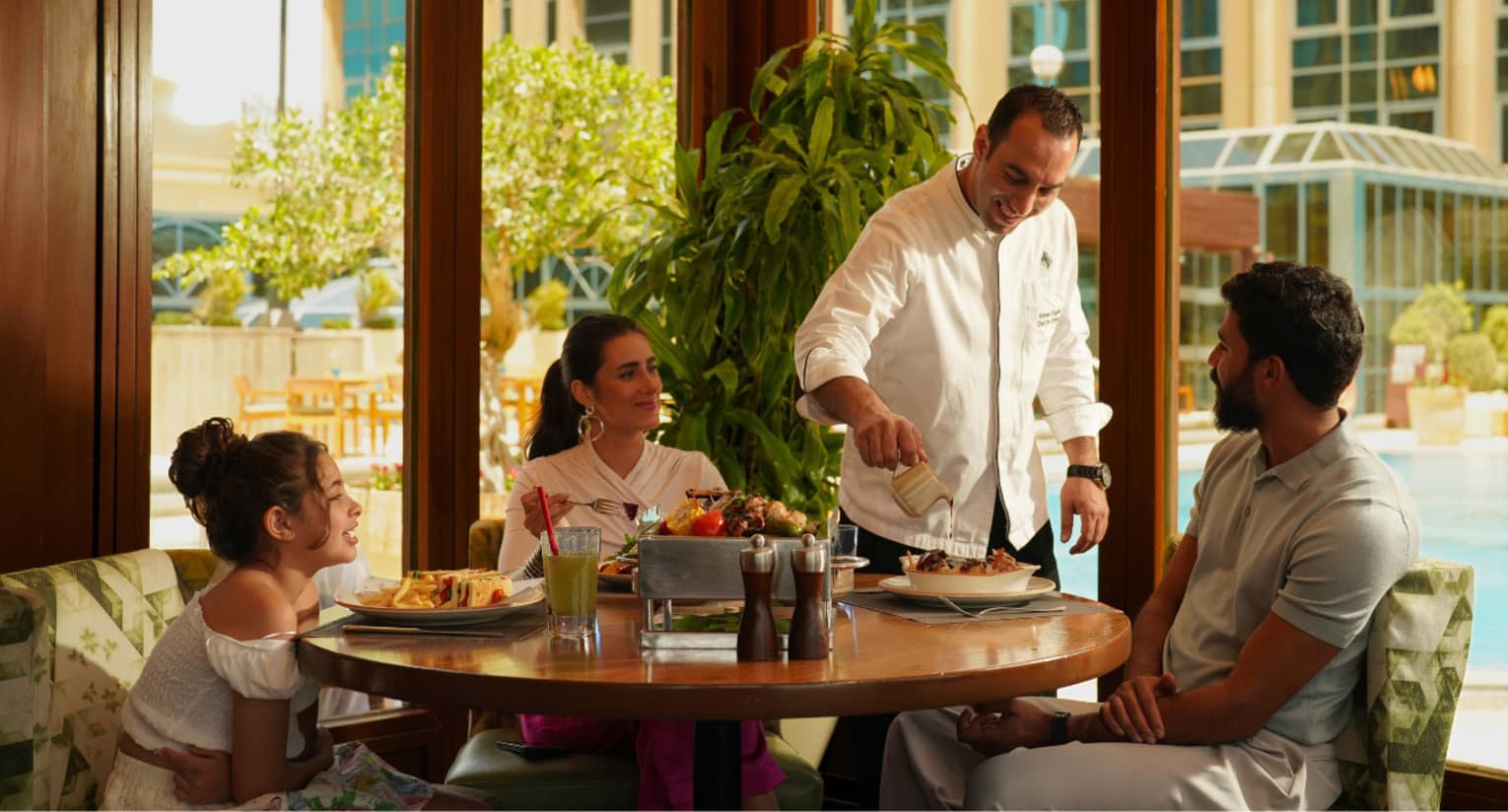 Family of three seated at round wooden table at poolside restaurant are served by chef in white coat