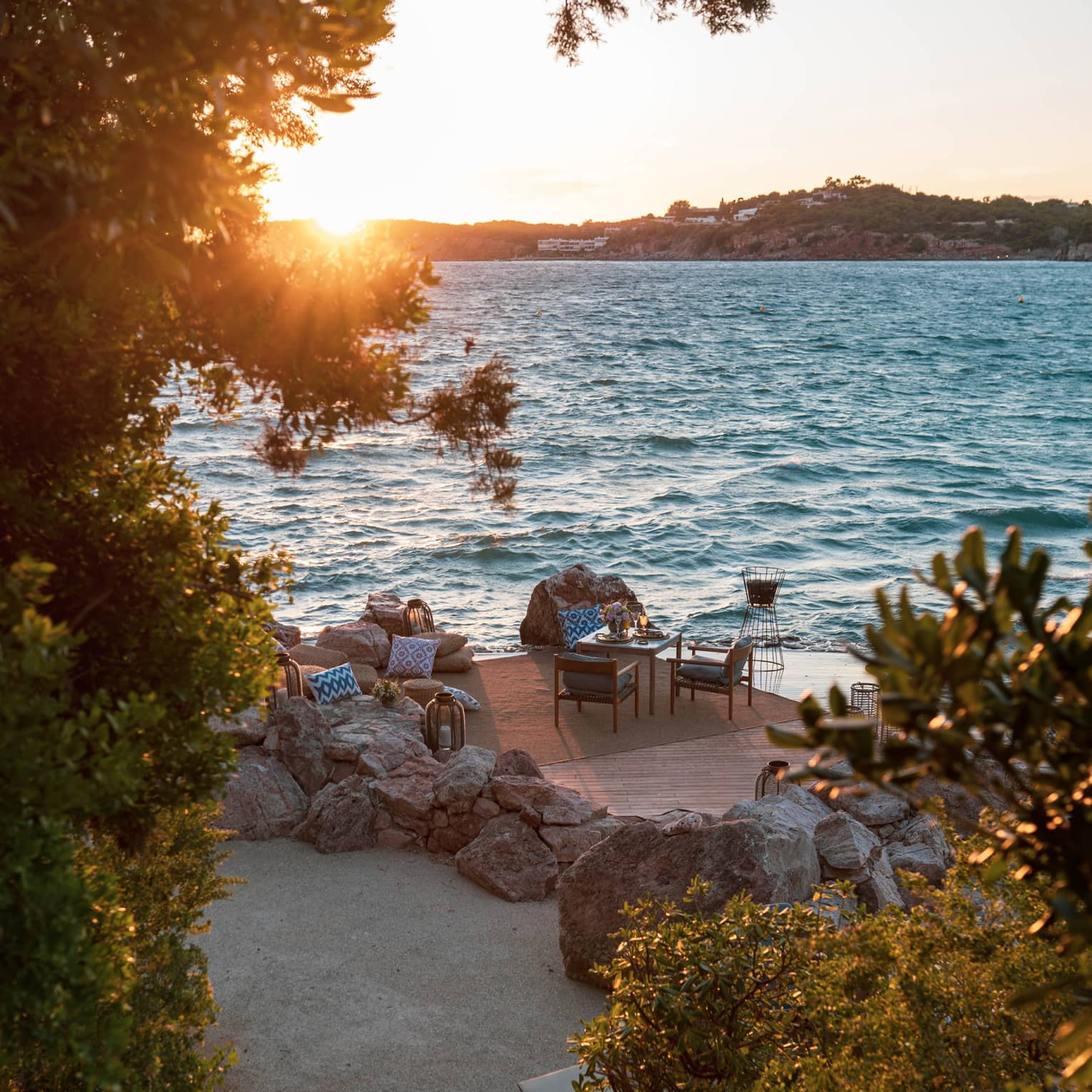 From tree-lined stairs, a furnished patio, including table set for two beside rippling water  and sun setting over a distant isle