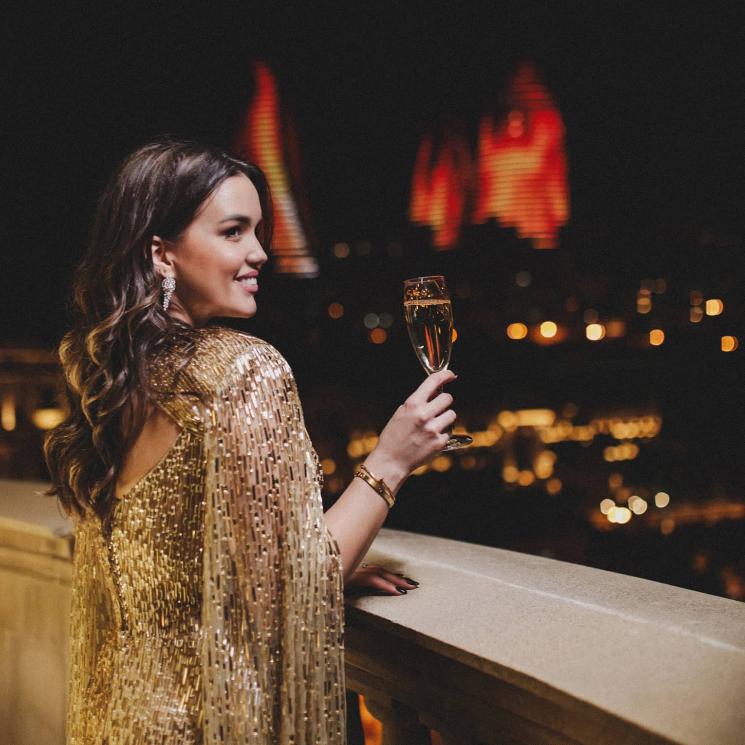 Woman with long brown hair in elegant gold dress holding a champagne flute on terrace at night