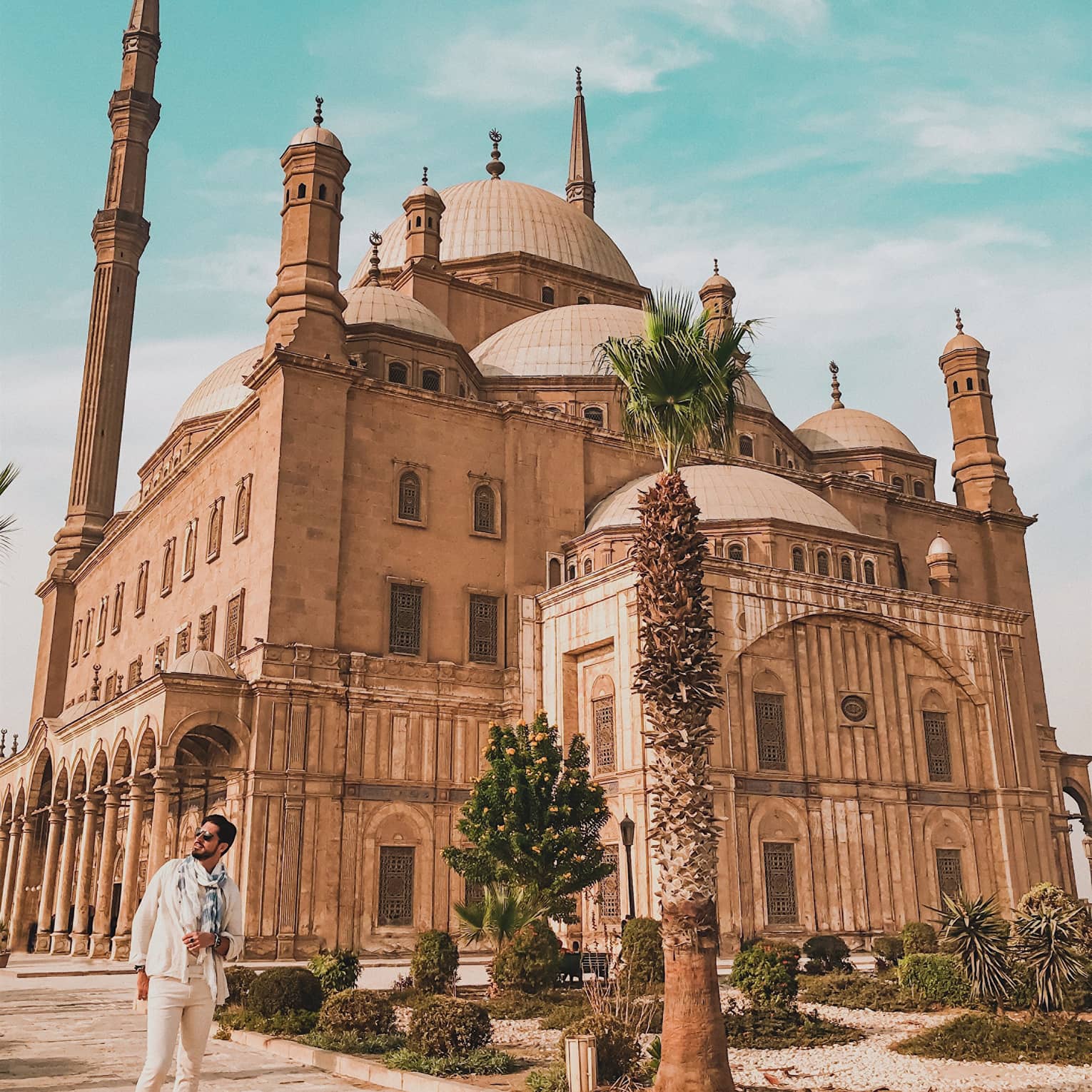 A tourist poses in front of the imposing Mosque of Muhammad Ali, minarets towering above the many domes of the building.