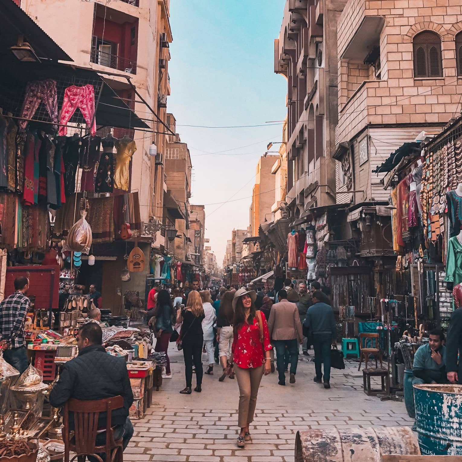 A smiling tourist strolls down a market alley, surrounded by varoius wares of the many merchants crowded between buildings. 