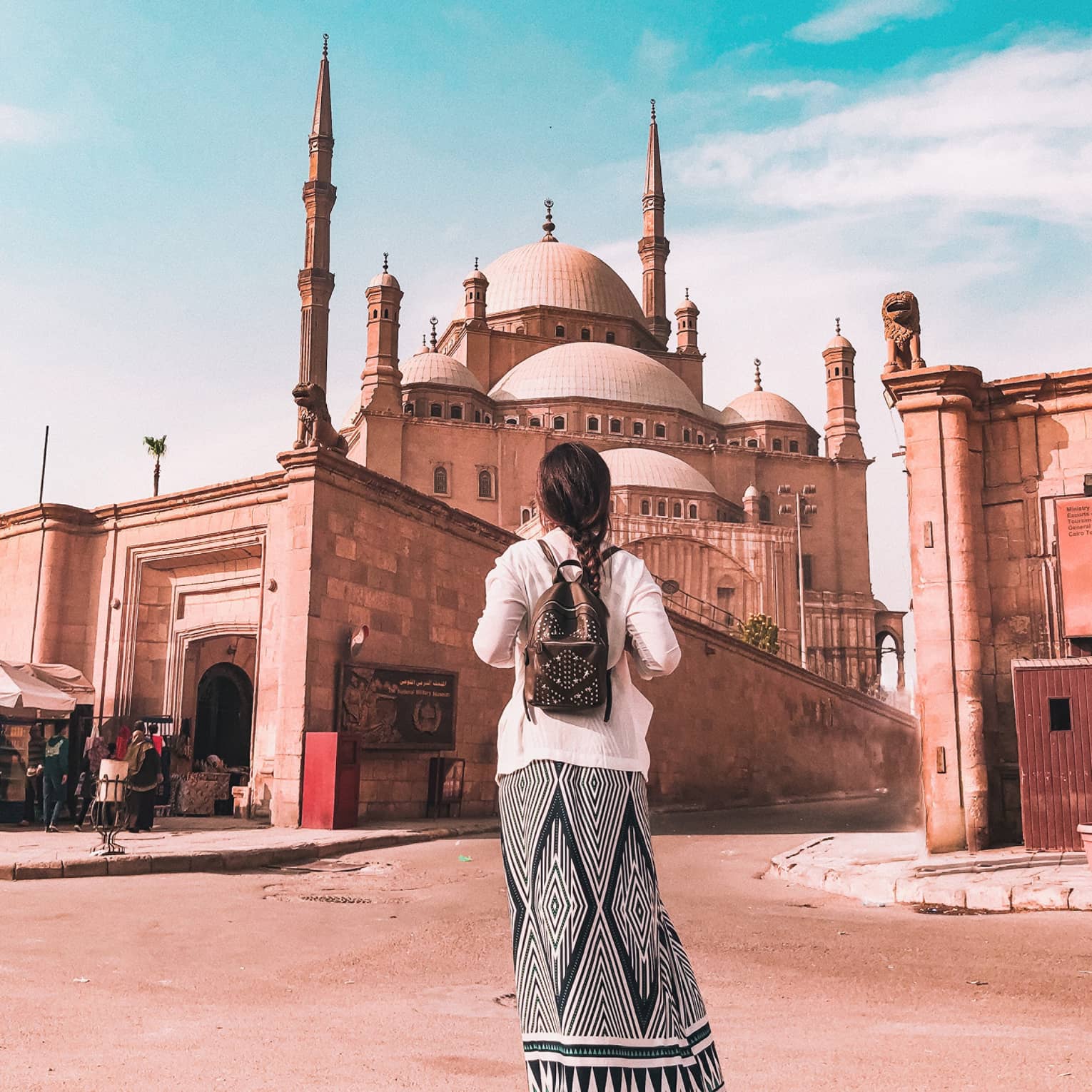 A tourist in a long skirt faces away, observing the Mosque of Muhammad Ali that towers over a wall surrounding it. 