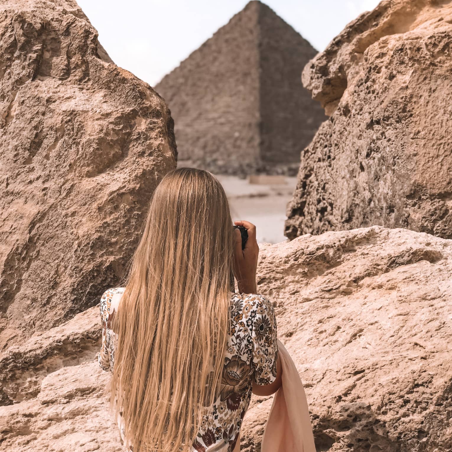 A tourist in a floral dress stands between two large rocks and aims her camera at an enormous pyramid beyond. 