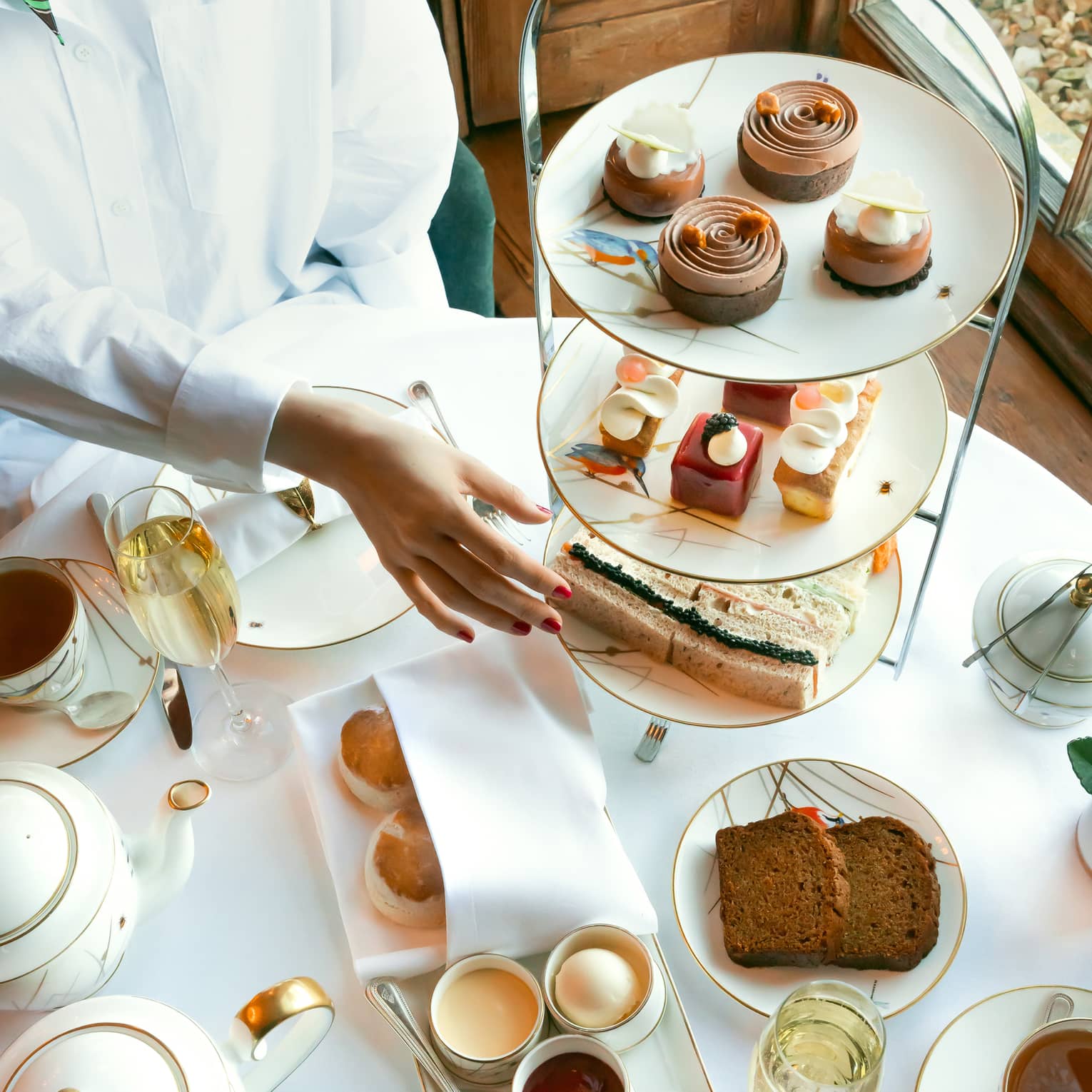 Person in white shirt reaches to classic afternoon tea stand filled with patisserie and mini sandwiches
