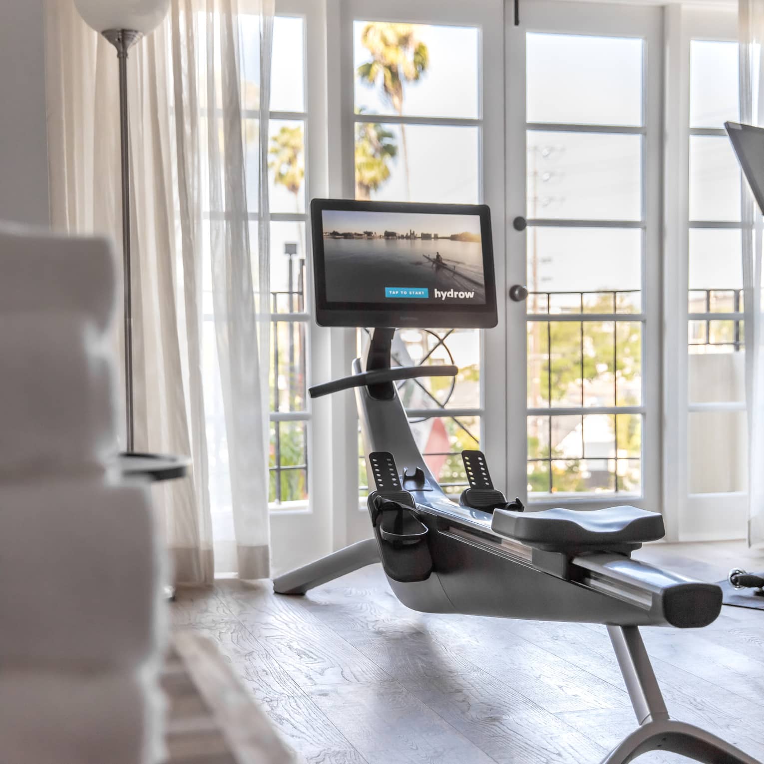 In a bright room, a rowing machine equipped with a screen faces two French doors, sheer white curtains pulled to the side.
