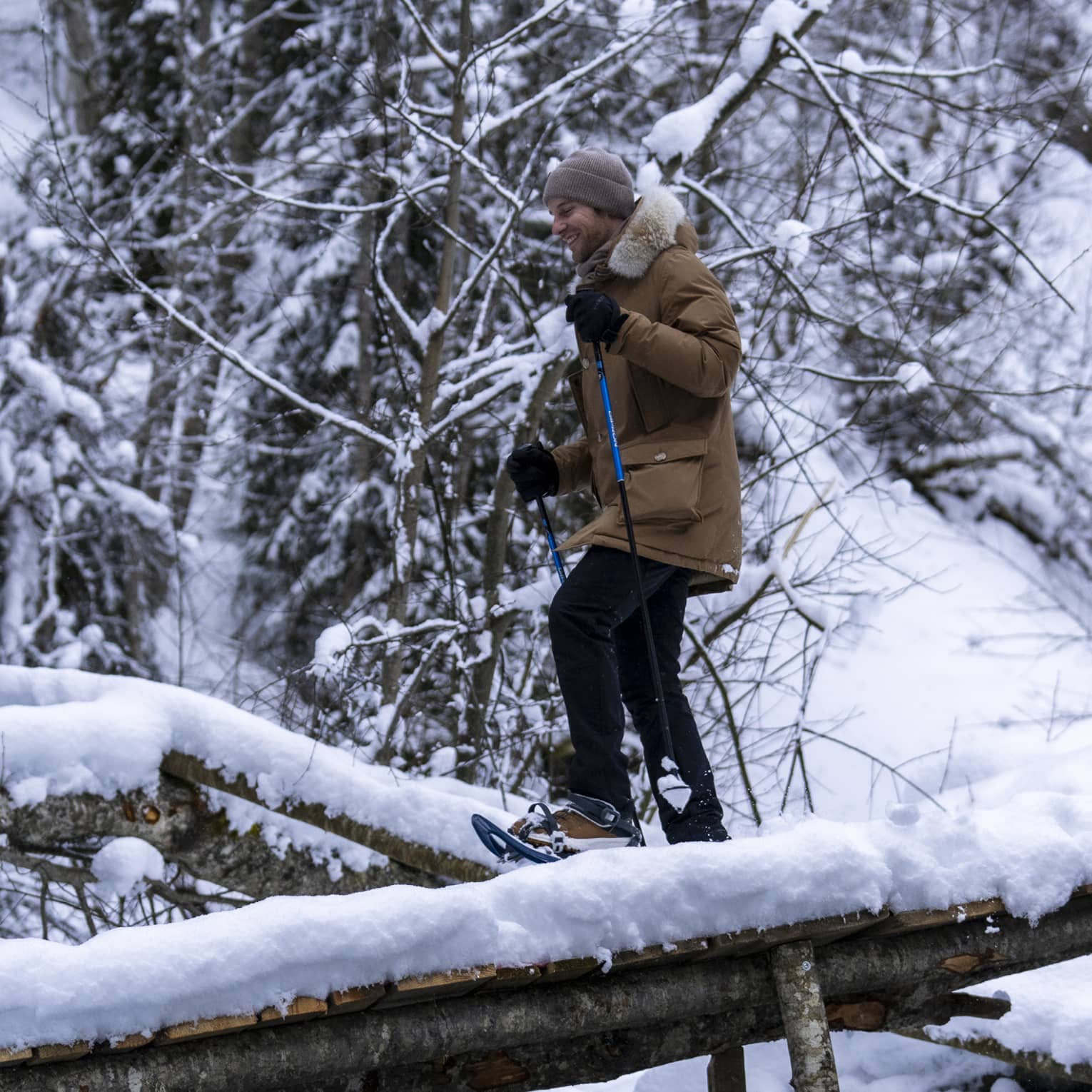A snowshoer in a knit cap, parka and gloves uses poles to cross fallen logs in a dense, snow-covered forest.