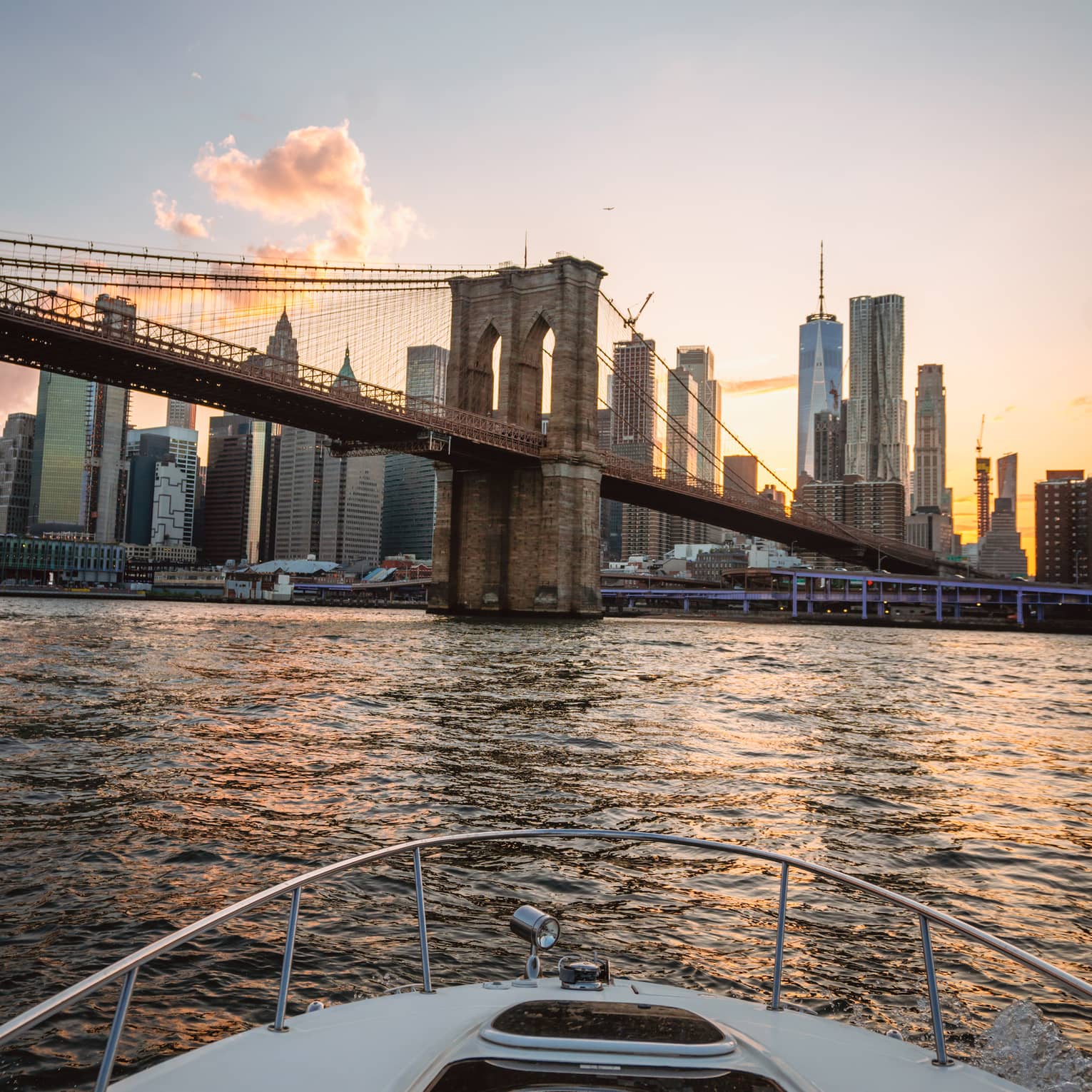 View from a boat of the Manhattan skyline behind the Brooklyn Bridge as a golden sunset reflects on the rippling water.