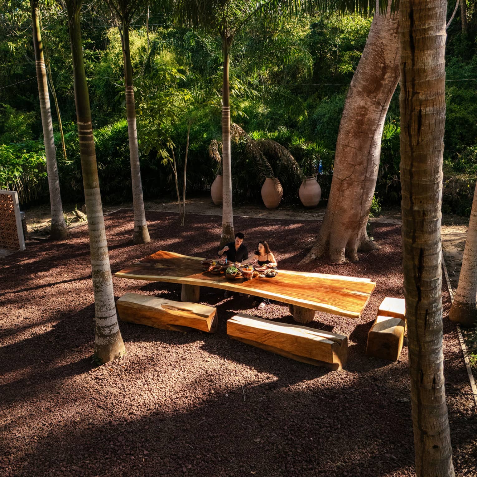 Two people sit at a long wood outdoor table with wood beam bench seats, all surrounded by palm trees.