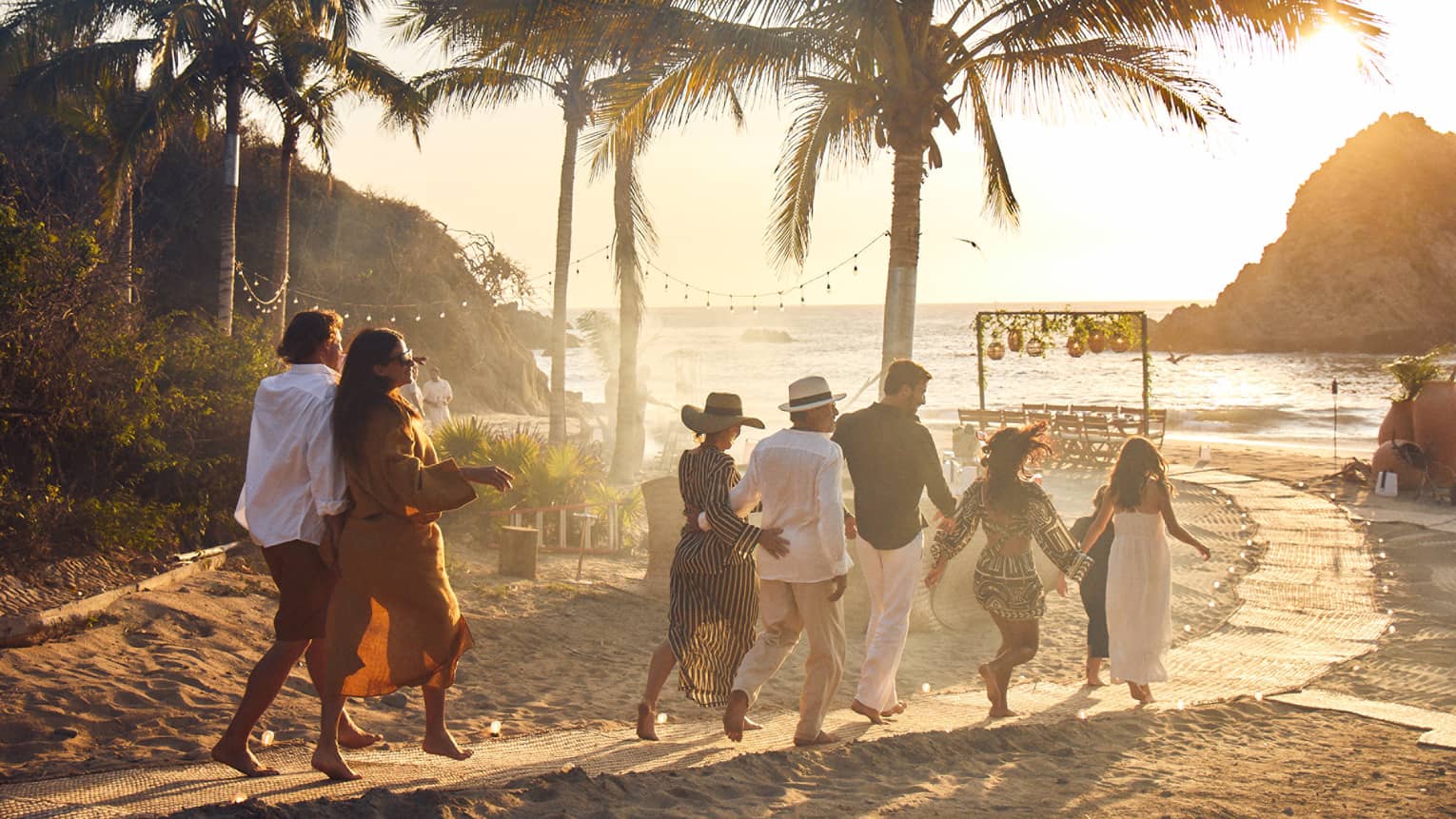 A group of adults walks down a palm-lined beach path towards a table by the ocean as the sun sets behind a rocky outcrop.