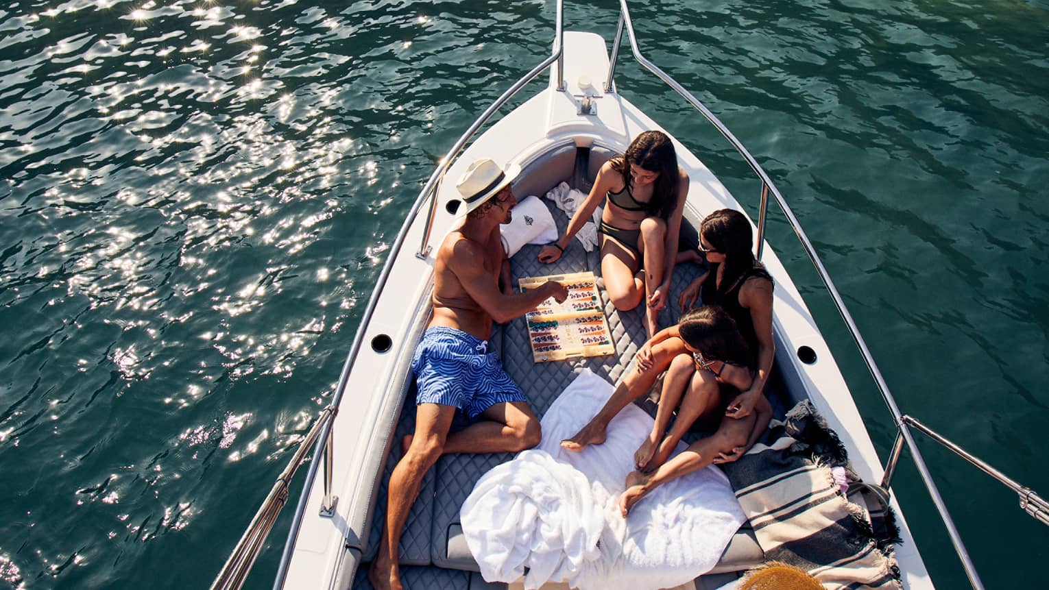 A family lounges in the bow of a luxurious boat amid towels and blankets, playing backgammon, the sun's glow on teal waters.