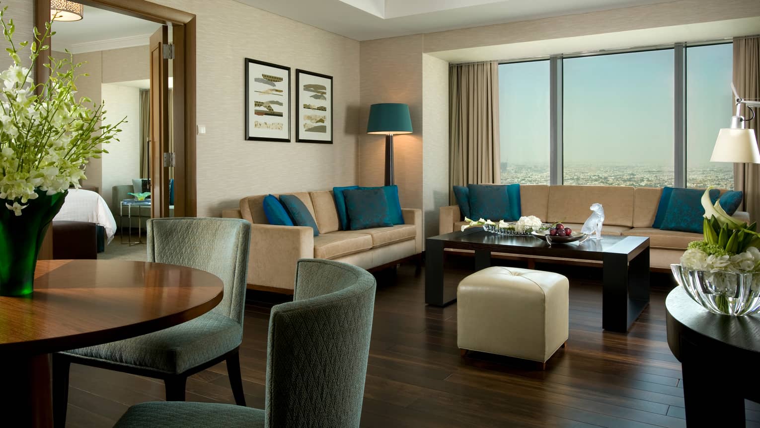 Large Superior Suite living room with two sofas with blue pillows by window, small dining table with plush chairs, flowers