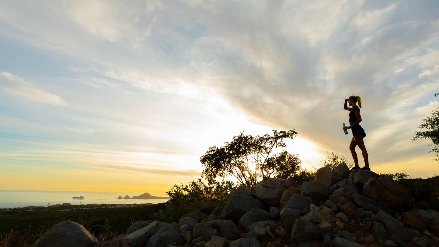 Silhouette of a woman standing on a rock with a water bottle in hand as the sun sets beyond the ocean