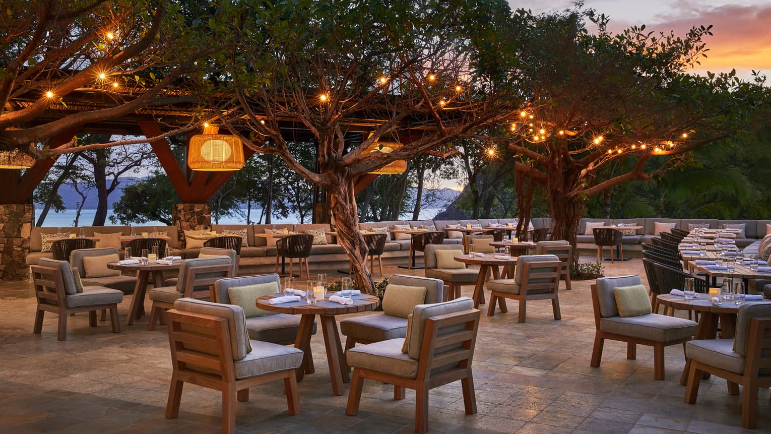 Patio tables, chairs with plush cushions on patio under trees with hanging lights, at sunset