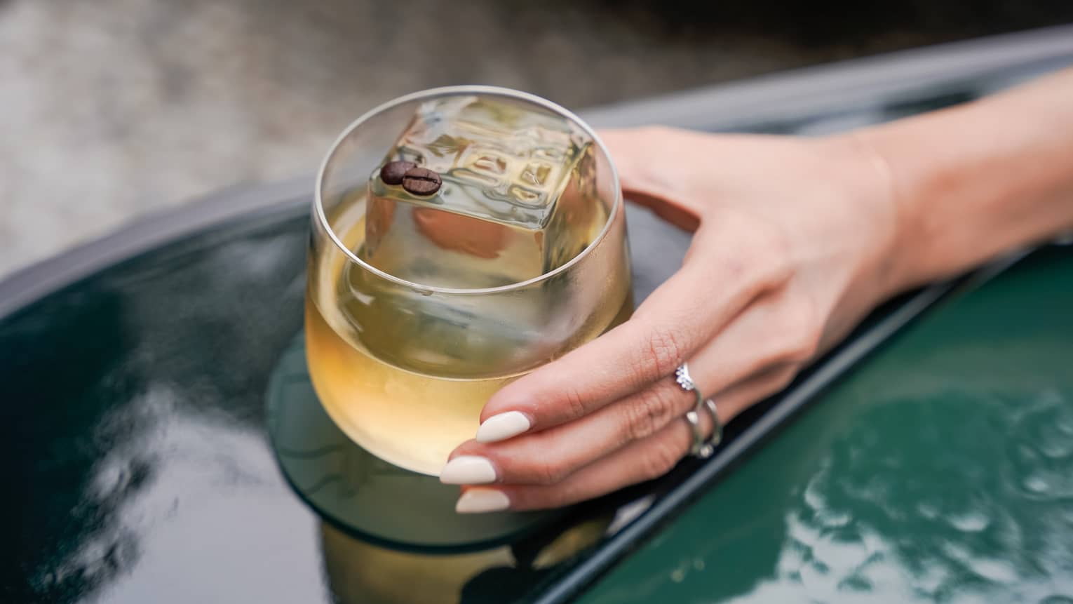 An outstretched hand holds a tan-hued cocktail in a rounded rocks glass