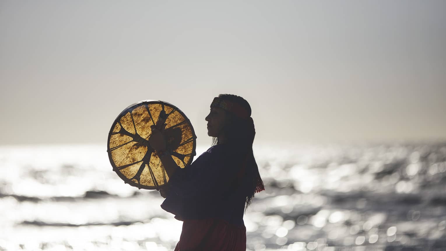 A woman holding a woven disc by the ocean.