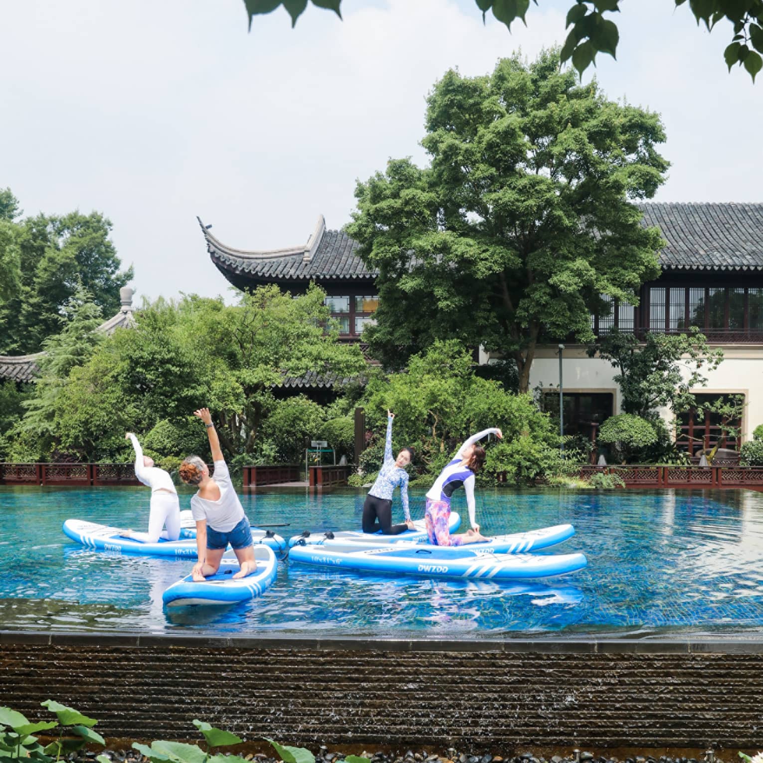 Four people kneel on paddleboards arranged like petals in a tranquil pool, one arm stretched skyward, amid lush foliage.