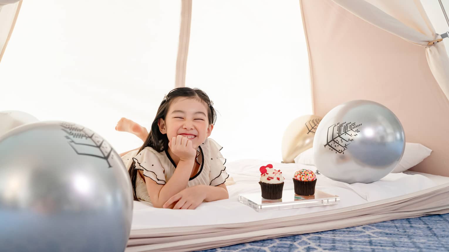 Smiling young girl in in-room kids? tent with gold and silver balloons and cupcakes
