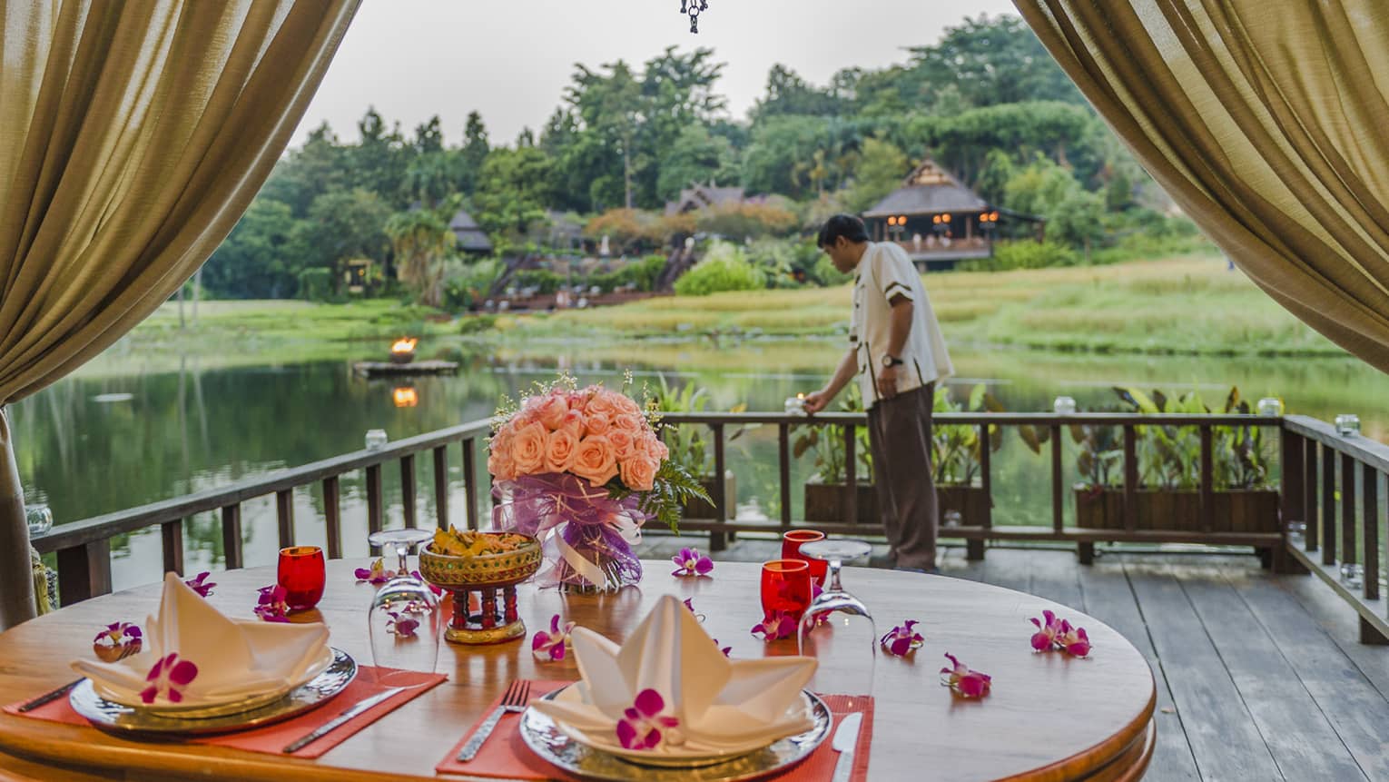 Private dining table with pink flowers, napkins on pavilion, hotel staff lights candle near water 