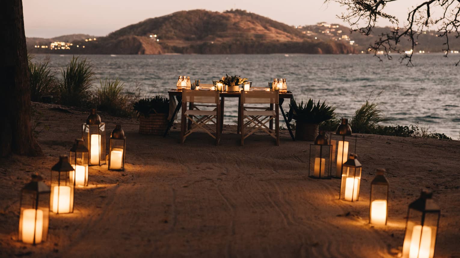 Lanterns line a path on the sand leading up to a table set for two overlooking the ocean