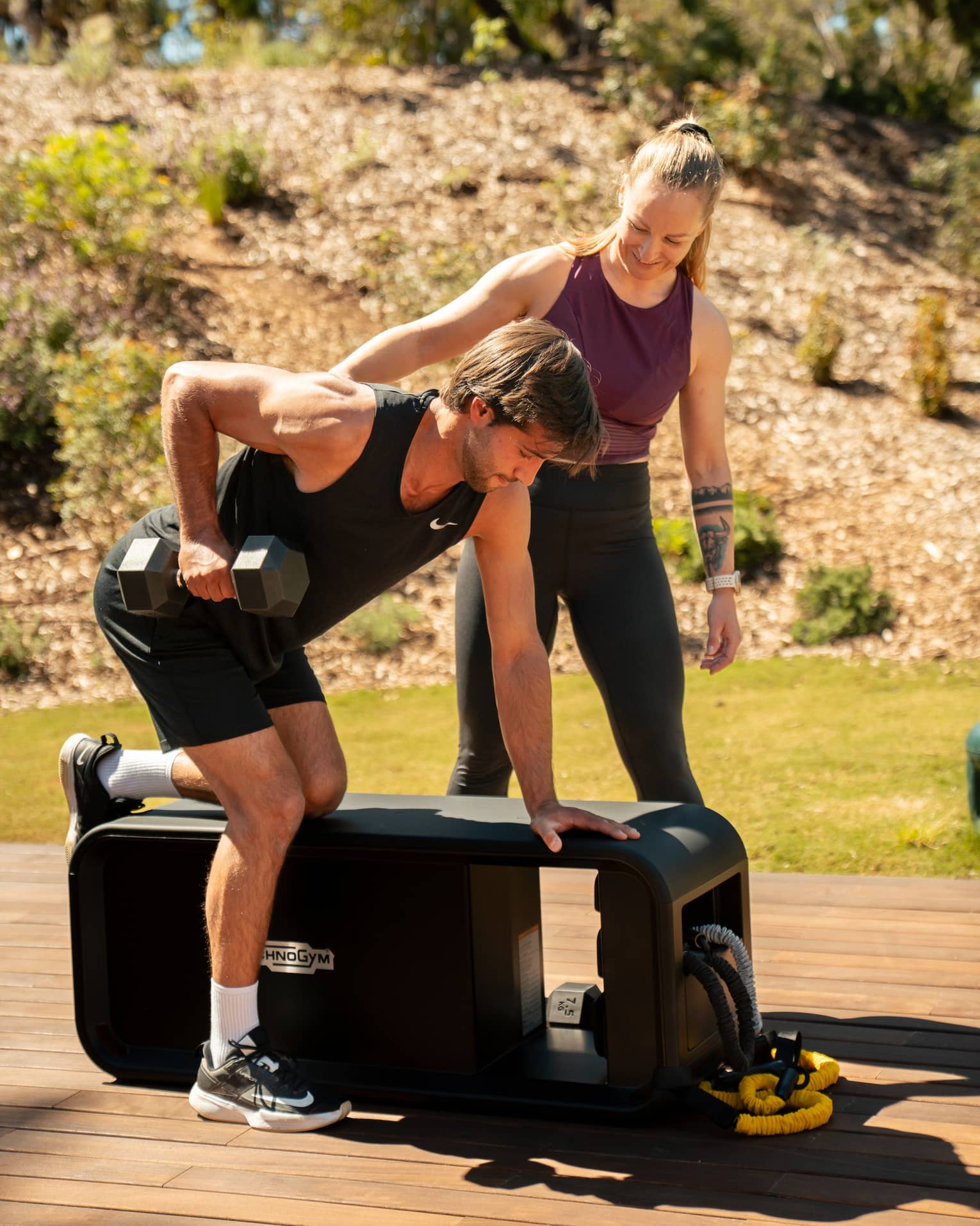 Two people exercise with weights at a workout bench on a wood deck in an outdoor setting.