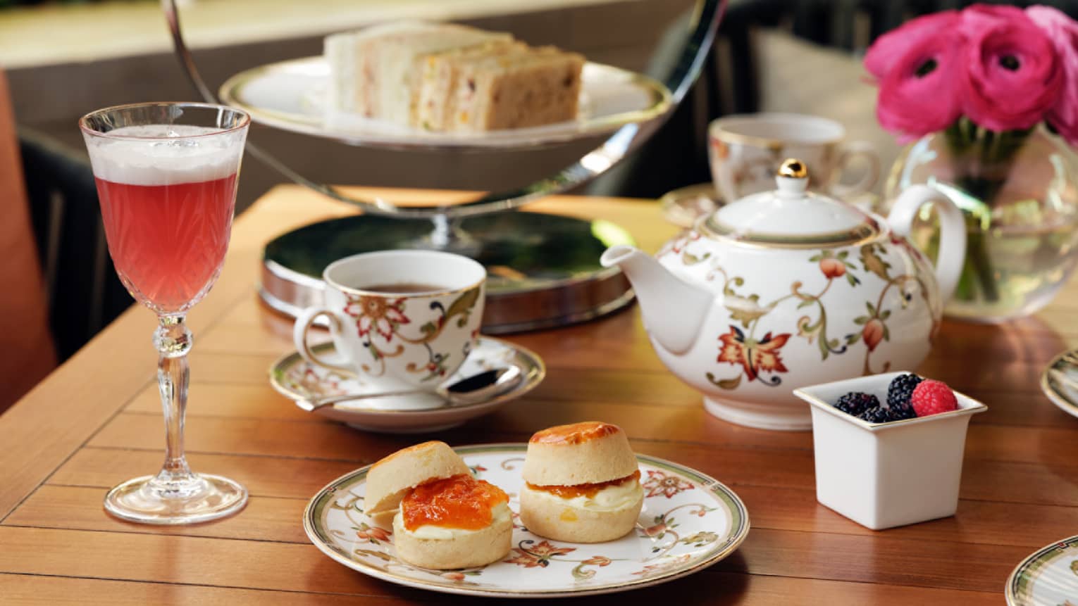A floral tea set next to scones and a glass of burgundy liquid.