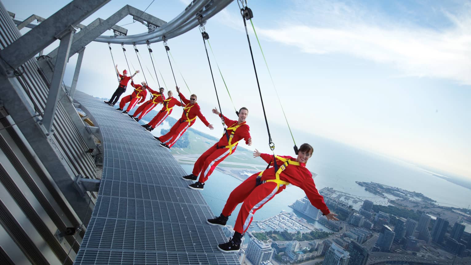People wearing red jumpsuits, harnesses lean back over edge of CN Tower high above city