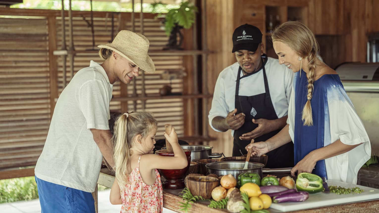 A chef instructs a guest stirring a pot, while a child uses a mortar and pestle across the counter as a third adult watches. 