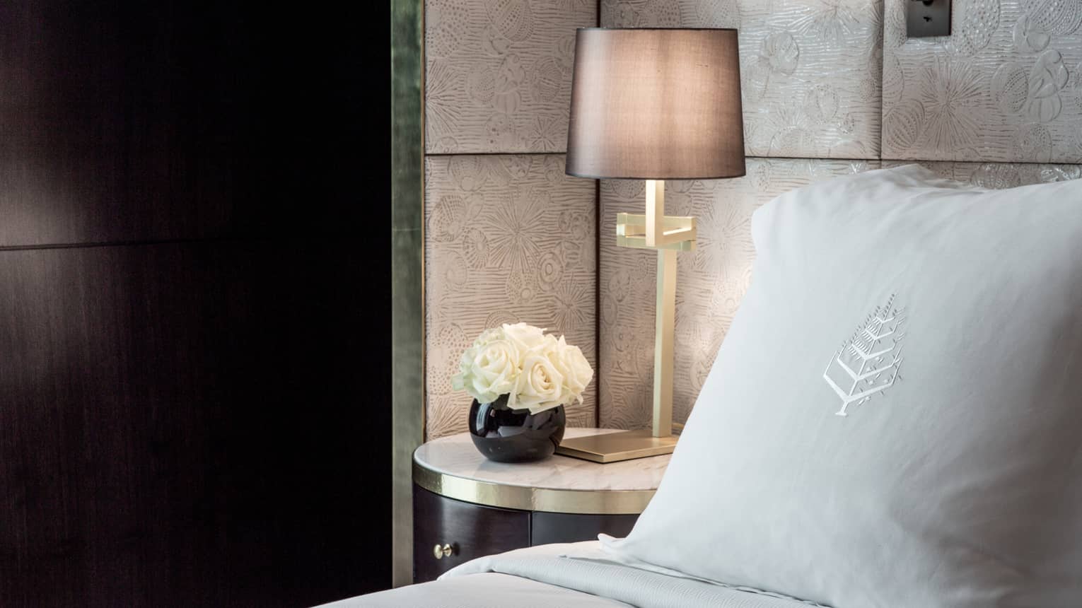 Premier room, close-up of pillow with Four Seasons logo, white roses in round black vase and bedside reading lamp