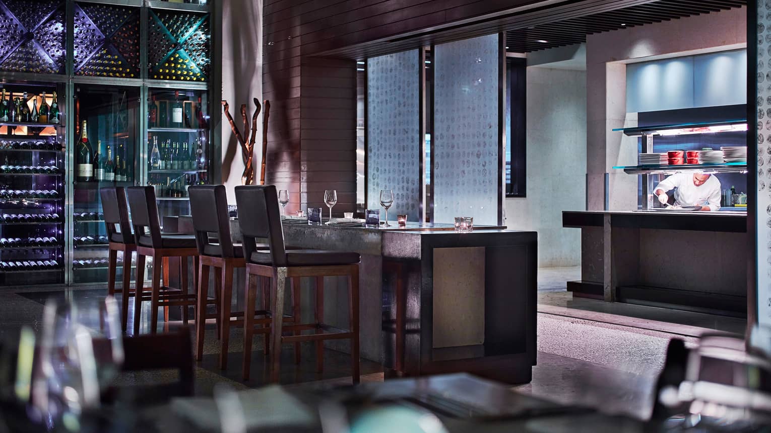 Beau Champ dimly-lit lounge with four stools at cement bar, chef in background