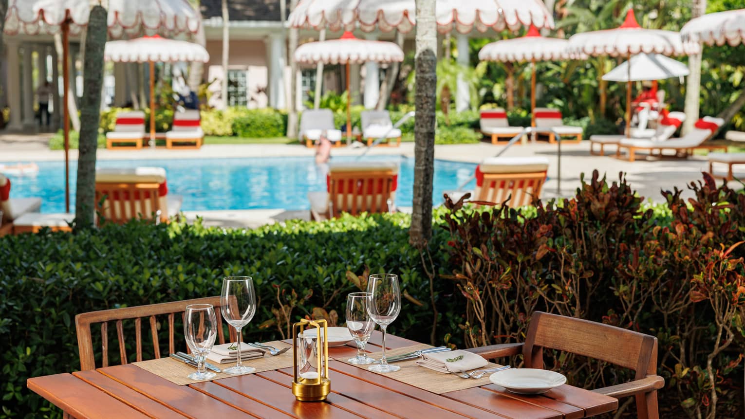 Wooden dining table topped with cutlery and glasses in front of an outdoor pool surrounded by white scalloped umbrellas and lounge chairs