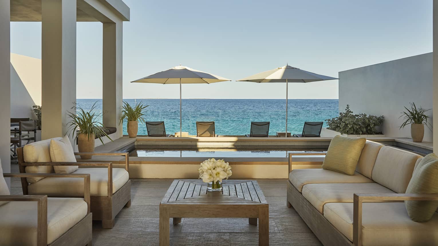 Outdoor patio living room with sofas, armchairs with white cushions, plunge pool, patio chairs and umbrellas, ocean