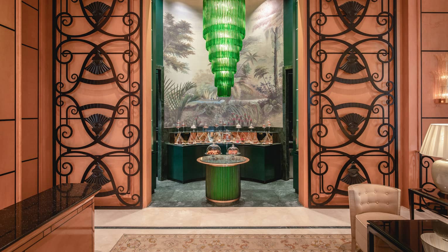 View through an ornate orange doorway of a tiered green chandelier hanging above tables of desserts covered by glass domes.
